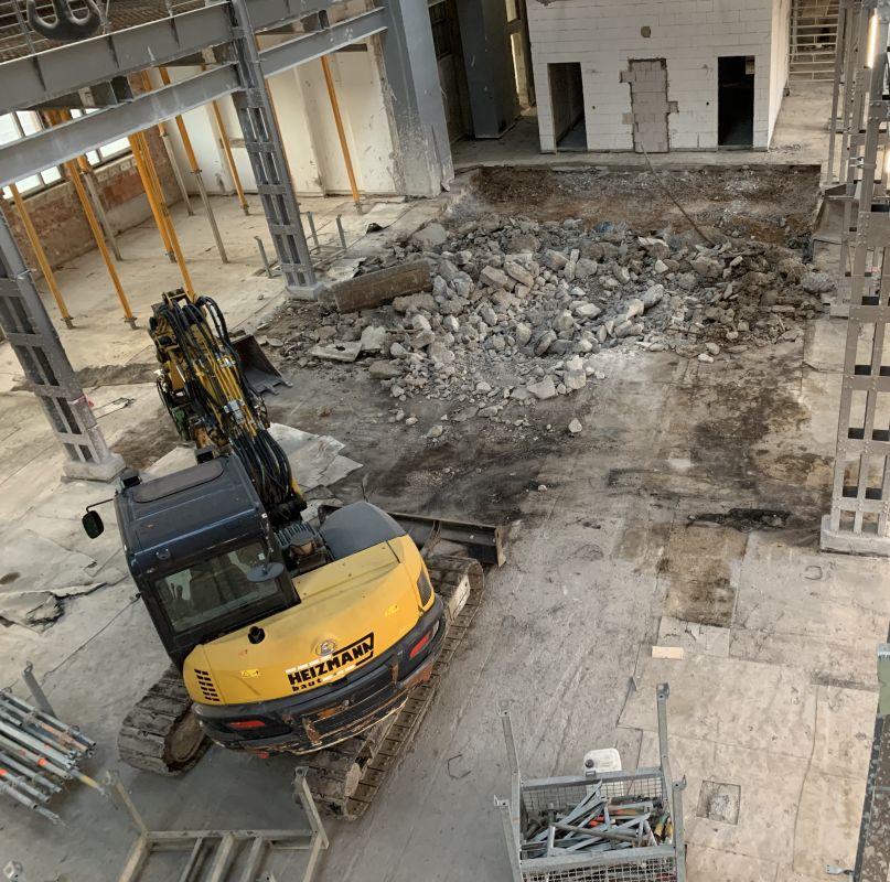 Eine Baustelle eines Bauunternehmens mit einem gelben Bagger und Schutthaufen in einem teilweise abgerissenen Gebäude.