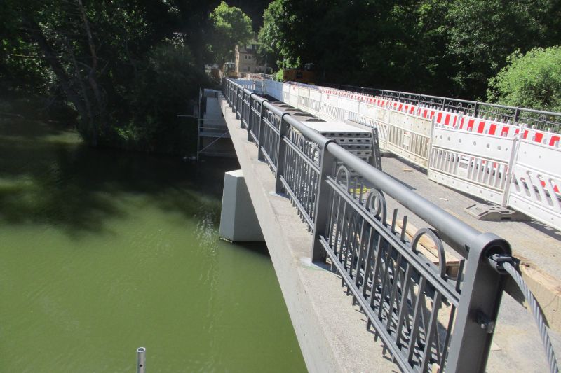 Im Bau befindliche Brücke über einen Fluss, mit Barrieren und Geländer; im Hintergrund Bäume und Laub. Das für die Hoch- und Tiefbauarbeiten verantwortliche Bauunternehmen sorgt für die termingerechte Fertigstellung des Projekts.