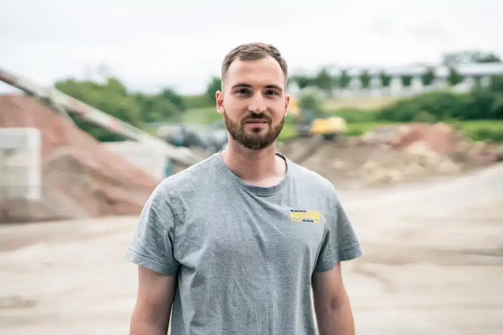 Ein b&auml;rtiger Mann in einem grauen T-Shirt steht im Freien auf einer Baustelle.